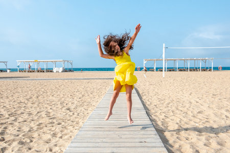 happy little curly girl in a yellow dress is jumping on the sandy beach on a sunny day. Fun holidays. Empty beachの写真素材