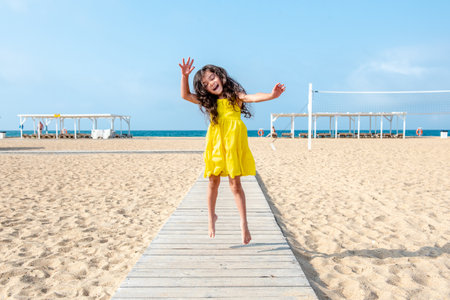 happy little curly girl in a yellow dress is jumping on the sandy beach on a sunny day. Fun holidays. Empty beachの写真素材