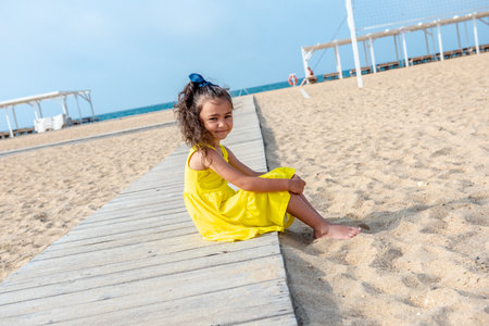 little curly girl in a yellow dress sits on a sandy deserted beach on a sunny day. Fun holidays. Empty beach. Summer fun. Self-isolationの写真素材