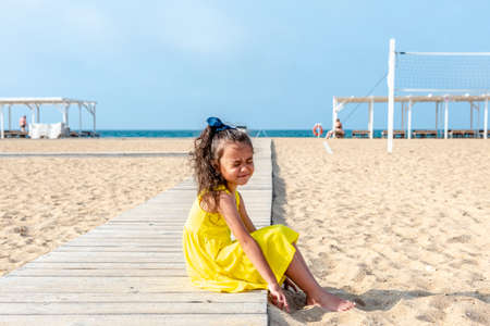 little curly girl in a yellow dress sits on a sandy deserted beach on a sunny day. Fun holidays. Empty beach. Summer fun. Self-isolationの写真素材