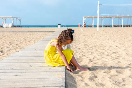 little curly girl in a yellow dress sits on a sandy deserted beach on a sunny day. Fun holidays. Empty beach. Summer fun. Self-isolationの写真素材