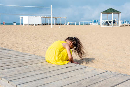 little curly girl in a yellow dress sits on a sandy deserted beach on a sunny day. Fun holidays. Empty beach. Summer fun. Self-isolationの写真素材