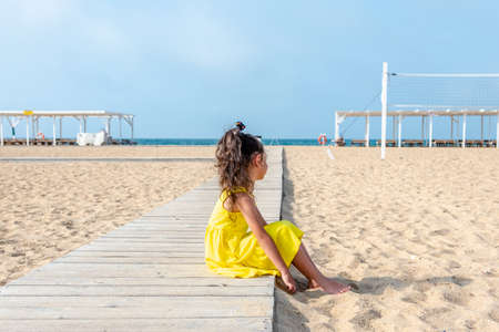 little curly girl in a yellow dress sits on a sandy deserted beach on a sunny day. Fun holidays. Empty beach. Summer fun. Self-isolationの写真素材