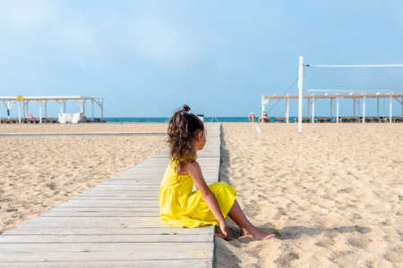 little curly girl in a yellow dress sits on a sandy deserted beach on a sunny day. Fun holidays. Empty beach. Summer fun. Self-isolationの写真素材