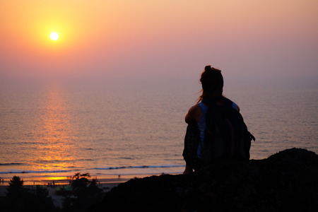 Female sitting on the edge of the hill. Silhouette of the girl with a backpack on the sunset.の写真素材