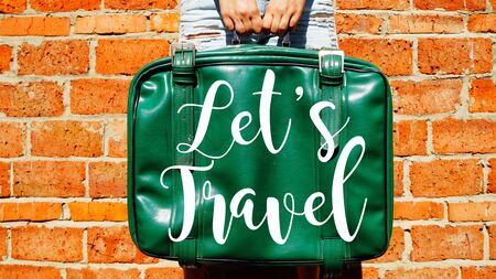 The girl is holding an old suitcase. On a suitcase written - Let's Travel. Red brick wall on the...
