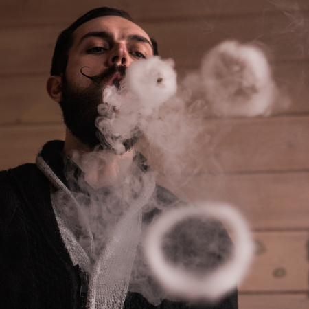 Young Man with Beard and Mustages Vaping an Electronic Cigarette. Vaper Hipster Smoke Vaporizer and Exhals Smoke Rings.の写真素材