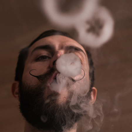 Young Man with Beard and Mustages Vaping an Electronic Cigarette. Vaper Hipster Smoke Vaporizer and Exhals Smoke Rings.の写真素材