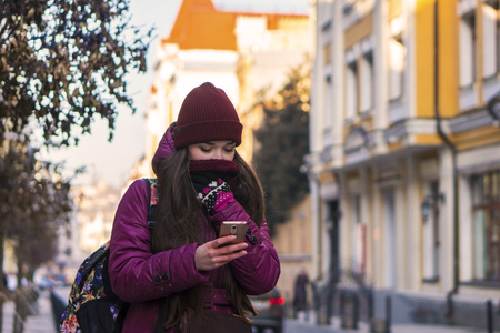 Pretty Brunette Girl Wearing Purple Winter Coat, Hat and Scarf, Walking by European Street at Winter, Using Her Smartphone and Making Selfieの写真素材