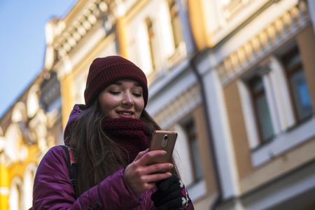 Pretty Brunette Girl Wearing Purple Winter Coat, Hat and Scarf, Walking by European Street at Winter, Using Her Smartphone and Making Selfieの写真素材
