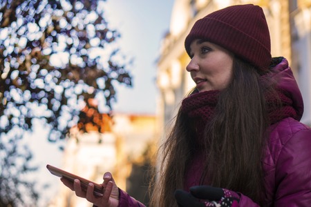 Pretty Brunette Girl Wearing Purple Winter Coat, Hat and Scarf, Walking by European Street at Winter, Using Her Smartphone and Making Selfieの写真素材