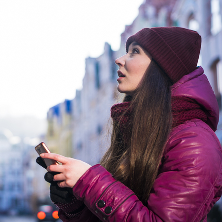 Pretty Brunette Girl Wearing Purple Winter Coat, Hat and Scarf, Walking by European Street at Winter, make Photos on Her Smartphone and Making Selfieの写真素材