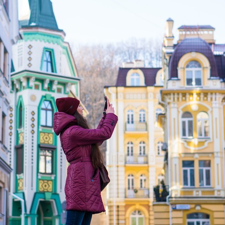 Pretty Brunette Girl Wearing Purple Winter Coat, Hat and Scarf, Walking by European Street at Winter, make Photos on Her Smartphone and Making Selfieの写真素材