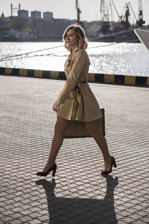 Attractive Young Blonde Woman in Trench with Vintage Suitcase on the Jacht Pierの写真素材