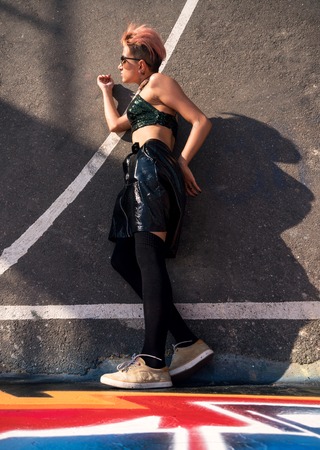 Stylish Girl at painted Basket Box. Street punk girl with pink dyed hair. Woman with piercing walk by wall at basketball court with graffiti paintings on wallsの写真素材