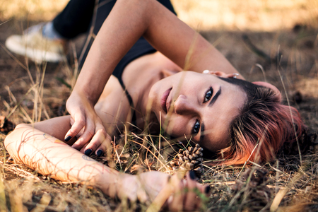Girl lay on Coniferous Litter. Young Pretty Fit Punk Girl Joying in Black Top with Pink Hair in Pine Forest at Sunset Timeの写真素材