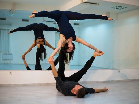 Young athletic couple in dark suits practicing acroyoga in studio with mirrors. Balancing in pairの写真素材