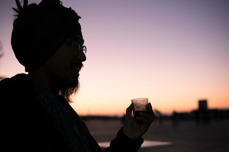 Young Asian Bearded Tourist man with Dreadlocks and scarf on head Drinking Morroccan coffee in Essaouira, Morocco at the evening sunset timeの写真素材