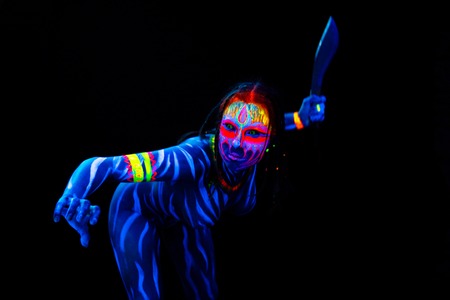Portrait of Young naked bodyarted woman in blue glowing ultraviolet paint with big machete knife. Agressive avatar warrior woman amazon with pigtails hairstyleの写真素材