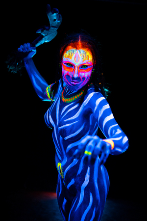 Portrait of Young naked bodyarted woman in blue glowing ultraviolet paint with tomahawk ancient prehistorical weapon. Agressive avatar warrior amazon with pigtails hairstyleの写真素材