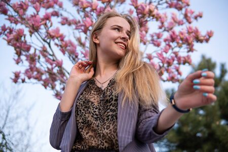 Portrait of Beautiful blonde girl posing with blooming Magnolia tree branches with pink flowers on...