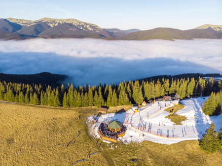 Aerial panorama of Bukovel Ski Resort mountain Dovha, Particularly covered with a Man-made Snow. Low clouds hang around the mountain and People on Skies and Snowdoards Are enjoying Sunset in Top View Panorama Restaurant.の写真素材