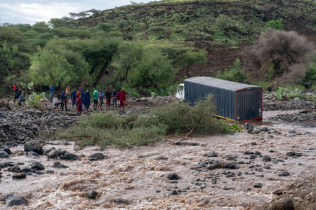 TANZANIA, ARUSHA - JAN 2020: Maasai people Standing on the Shore Deciding what to do while Heavy Cargo Truck Stuck in the River. Ford at African Mainland Roads. East Africaのeditorial素材