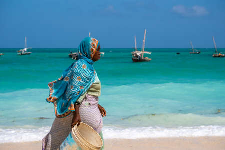 NUNGWI, ZANZIBAR - JAN 20, 2019: local woman in Bright Cloth walking by the Indian ocean Beach with a Bucket for Fish Nungwi village, Zanzibar island, Tanzaniaのeditorial素材