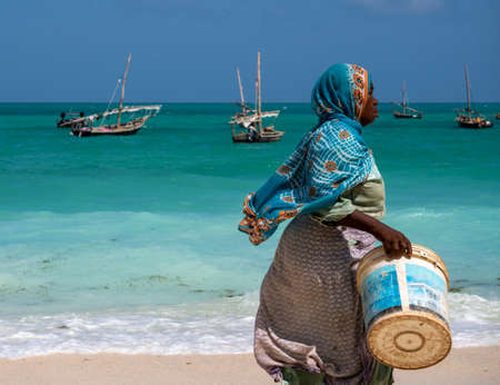 NUNGWI, ZANZIBAR - JAN 20, 2019: local woman in Bright Cloth walking by the Indian ocean Beach with a Bucket for Fish Nungwi village, Zanzibar island, Tanzaniaのeditorial素材
