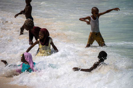 Nungwi, Zanzibar - December 2019: Boys and Girls in Turquoise Water. African children playing on the beach Ocean Surf Waves at Sunny Day at Nungwi village in northern Zanzibar, Tanzaniaのeditorial素材