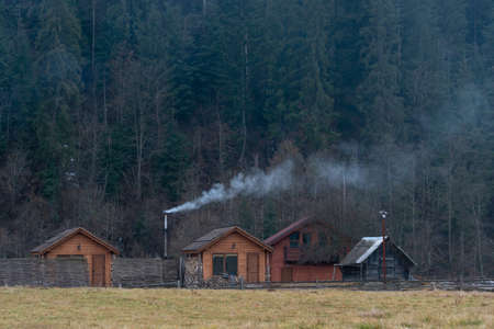 White smoke from the chimneys of Accurate Wooden houses in a mountain village in Autumn on a background of forest and mountainsの写真素材
