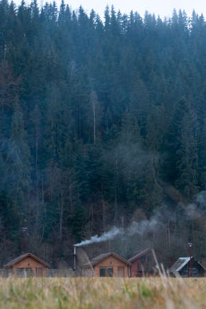 White smoke from the chimneys of Accurate Wooden houses in a mountain village in Autumn on a background of forest and mountainsの写真素材