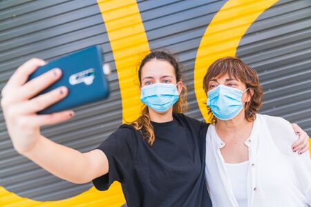 Mother and daughter making a selfie in the street protected by a mask. Family protected with a mask. Family selfie with mask. Selfie with mask. Human life in Social distance. New normal.の写真素材
