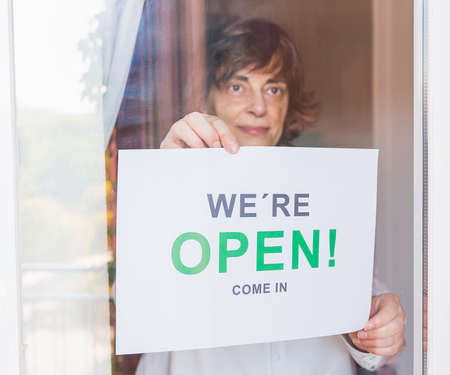 Business owner with a face mask hanging the open sign in her restaurant. Woman with a face mask...