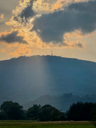 A view of the sun setting between the clouds over a forested mountain with an antenna on its top.の写真素材