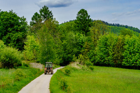 An old tractor drives along a winding rural road surrounded by green meadows, with trees, distant mountains, and cloudy sky.の写真素材