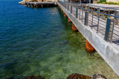 Angular jetty stretches above transparent shallows revealing algae and rocks beside deeper blue harbor.の写真素材