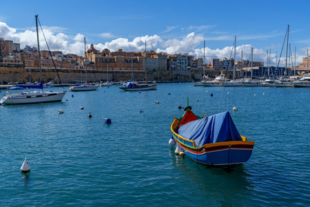 Colorful covered luzzu floats among buoys with yachts and limestone waterfront under bright blue sky.の写真素材