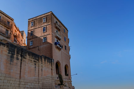 Sunlit masonry tower rises above high retaining wall, showing small windows, balconies and clean blue sky.の写真素材