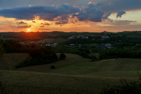 Golden sun sets behind village, colorful clouds, peaceful Tuscan landscape, hills and fields glowing.の写真素材