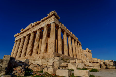 Striking low-angle shot of the Parthenon on the Acropolis, displaying its massive stone columns, partially ruined structure, and scattered stones against a deep blue sky.の写真素材