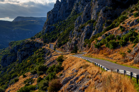 Mountain road with white guardrail snakes along a steep rocky slope covered in dry grass and shrubs, high above a shadowed gorge under a moody sky.の写真素材