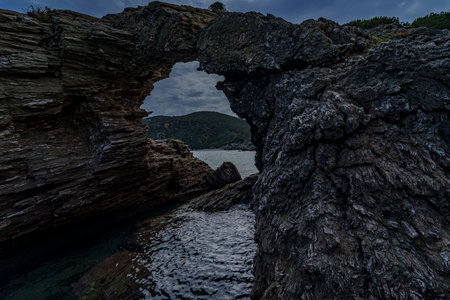 Wave-carved rock arch frames distant headland beneath a brooding, overcast evening sky.の写真素材