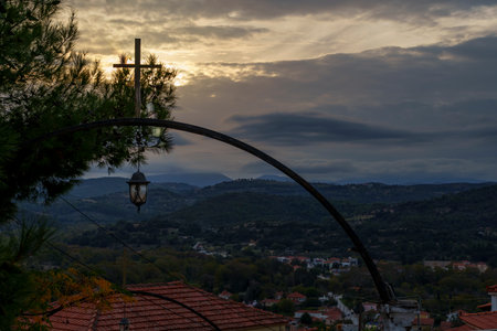 Silhouette of a metal arch with a cross and lantern overlooks a village nestled among dark hills under a moody sunset sky.の写真素材