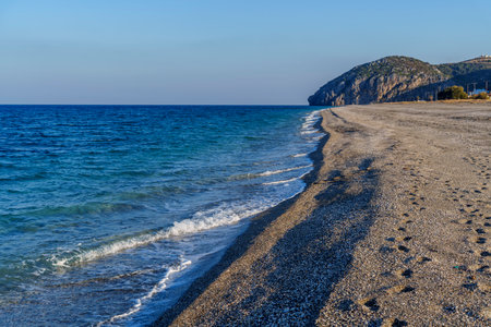 Empty pebble beach with gentle waves, clear blue sea and rocky headland at horizon.
