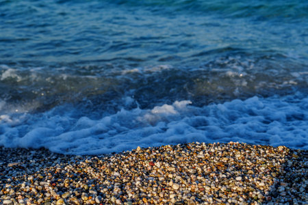 Close-up of small multicolored pebbles on a beach with soft foamy wave washing ashore and blue sea in background.の写真素材