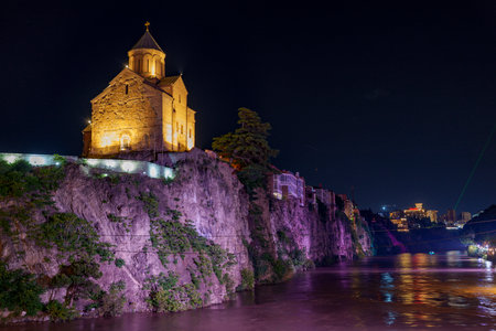 Stone church crowns steep rocky cliff, its warm lights reflecting in dark, slowly flowing river.
