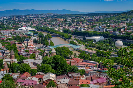 Wide cityscape shows Old Tbilisi rooftops, multiple churches, the Kura River with Peace Bridge and the modern tubular Rike Park concert halls against distant mountains.の写真素材