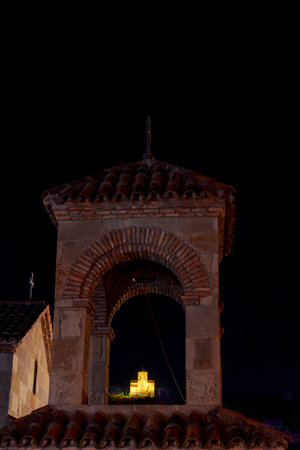 Rustic arched bell tower rises against dark sky, revealing distant glowing church through central...