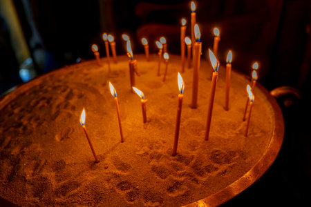 Thin devotional candles burning in a sand-filled tray inside dimly lit churchの写真素材
